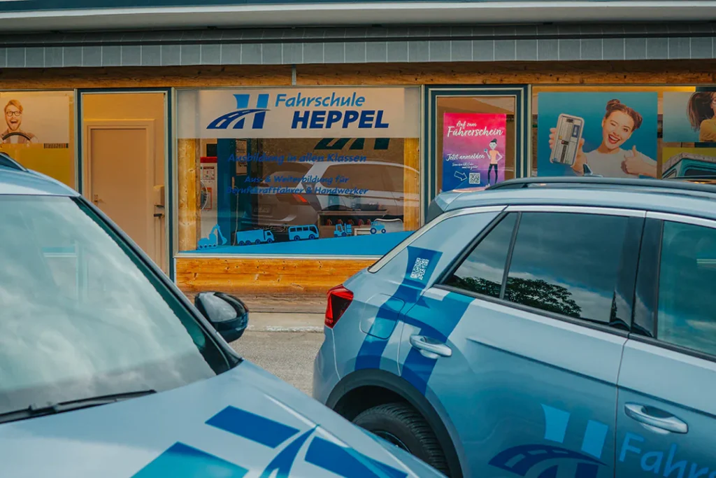 Fahrschule Heppel in Kiefersfelden von außen mit zwei Fahrschulautos vor dem Schaufenster.